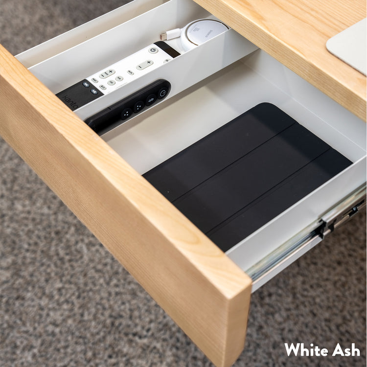 Minimal under-desk drawer in white ash wood, open to reveal white interior with tablet and remotes.