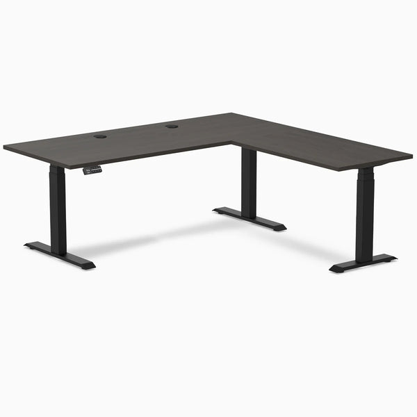 Burnished Wood L-shape laminate sit-stand desk with matte black legs and grommets.