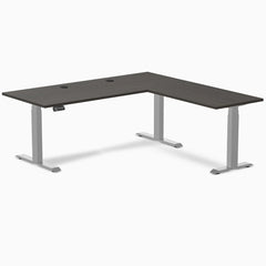 Burnished wood L-shape laminate sit-stand desk with gray frame and grommets.