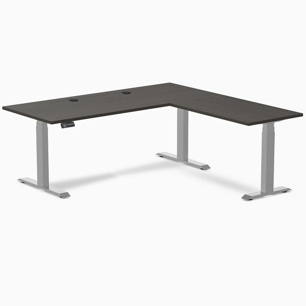 Burnished wood L-shape laminate sit-stand desk with gray frame and grommets.