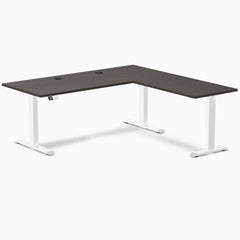 Burnished wood laminate L-shaped sit-stand desk with white adjustable legs and grommets.