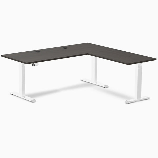 Burnished wood laminate L-shaped sit-stand desk with white adjustable legs and grommets.