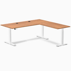 Prime Oak laminate L-shaped sit-stand desk with white adjustable legs and two black grommets.