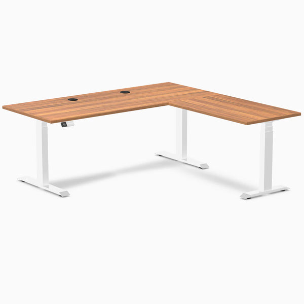 Prime Oak laminate L-shaped sit-stand desk with white adjustable legs and two black grommets.