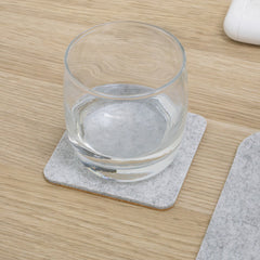 Square grey felt and cork coaster with a glass of water on a light wood desk.