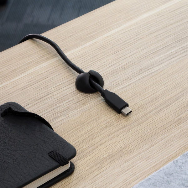 Black Desky Cable Organizer clip holding a USB-C cable on a light wood desk next to a black notebook.