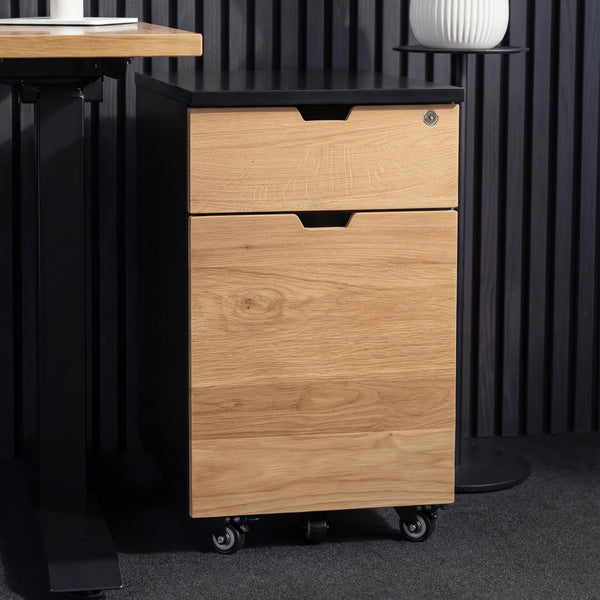 Desky wooden minimal filing cabinet with two oak drawers, black top, and casters, next to a desk.