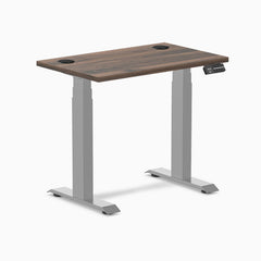 Desky Dual Mini sit-stand desk with Jarrah Legno top, gray legs, and two grommets.